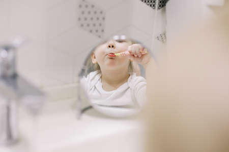 little funny girl brushing teeth and washing hands in the bathroom, reflection in the mirrorの写真素材