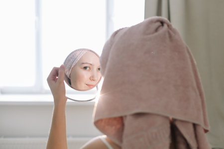 young woman with a towel on head looking in the mirror and apply cream on face skinの写真素材