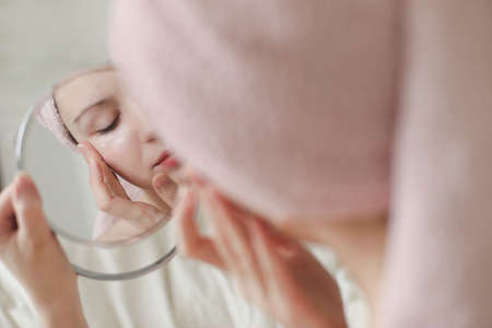young woman with a towel on head looking in the mirror and apply cream on face skinの写真素材