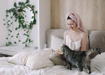 Young woman and her adorable cat reading book and resting in bed. Lazy weekend at home with loved pet concept. Close up, copy space, interior background. Love, togetherness and pets indoorsの写真素材