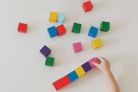 Top view on child hands playing and building with colorful wooden toy bricks on white wooden table....
