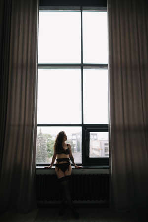 Gorgeous woman wearing black lingerie indoors. Portrait of a beautiful dreamy girl in a black underwear by the windowの写真素材