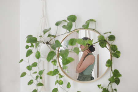 Young woman in apron, housewife or gardener takes care of green plant, watering flower pots in cozy room. Concept of gardening and hobbyの写真素材