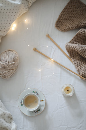 Cozy morning still life with a coffee cup, candle, needles and a woolen yarn in white bed. Knitting warm woolen sweater or scarf top view. Hygge lifestyle, cozy mood. Handicraft day concept.の写真素材