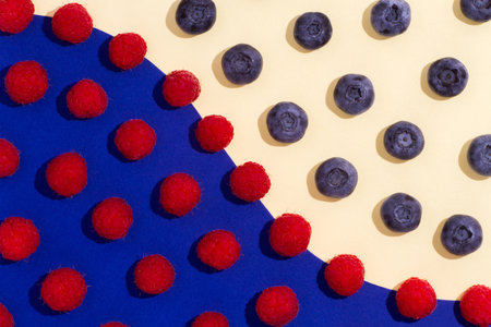 Blueberries and raspberry spaced in style flat lay on a double paper yellow-blue background.の写真素材