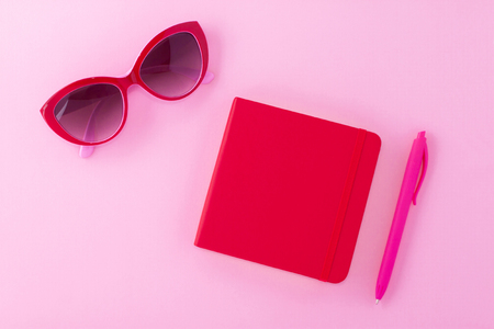 A red notepad on a light pink background with a sunglasses and bright fuchsia pen.の写真素材