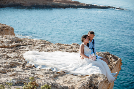 Beautiful smiling young bride and groom walking on the beach, kissing and having fun, wedding ceremony near the rocks and sea. Wedding ceremony on coast of Cyprusの写真素材
