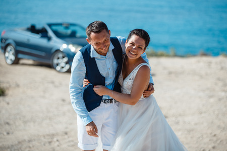 Beautiful smiling young bride and groom walking on the beach, kissing and having fun, wedding ceremony near the rocks and sea. Wedding ceremony on coast of Cyprusの写真素材