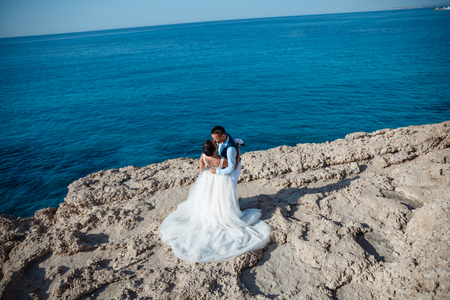 Beautiful smiling young bride and groom walking on the beach, kissing and having fun, wedding ceremony near the rocks and sea. Wedding ceremony on coast of Cyprusの写真素材