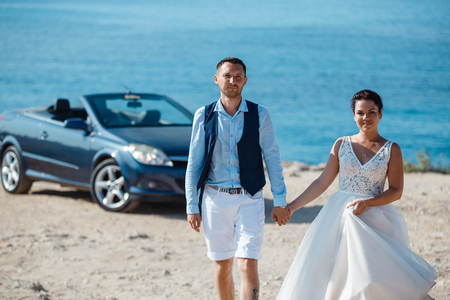 Beautiful smiling young bride and groom walking on the beach, kissing and having fun, wedding ceremony near the rocks and sea. Wedding ceremony on coast of Cyprusの写真素材