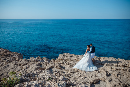 Beautiful smiling young bride and groom walking on the beach, kissing and having fun, wedding ceremony near the rocks and sea. Wedding ceremony on coast of Cyprusの写真素材