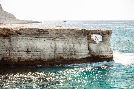 Beautiful smiling young bride and groom walking on the beach, kissing and having fun, wedding ceremony near the rocks and sea. Wedding ceremony on coast of Cyprusの写真素材
