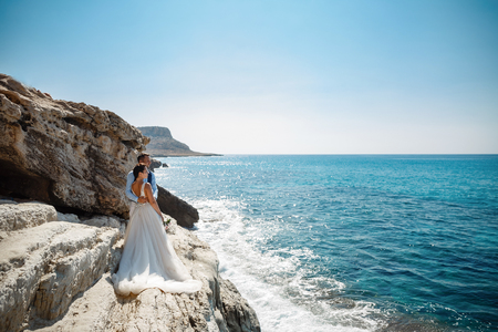 Beautiful smiling young bride and groom walking on the beach, kissing and having fun, wedding ceremony near the rocks and sea. Wedding ceremony on coast of Cyprusの写真素材