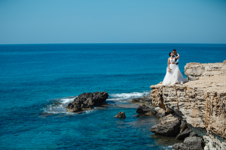 Beautiful smiling young bride and groom walking on the beach, kissing and having fun, wedding ceremony near the rocks and sea. Wedding ceremony on coast of Cyprusの写真素材