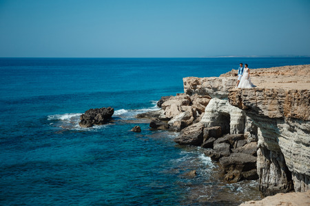 Beautiful smiling young bride and groom walking on the beach, kissing and having fun, wedding ceremony near the rocks and sea. Wedding ceremony on coast of Cyprusの写真素材