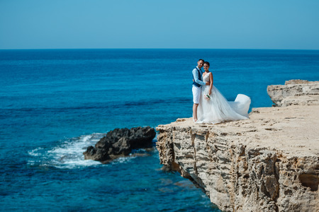 Beautiful smiling young bride and groom walking on the beach, kissing and having fun, wedding ceremony near the rocks and sea. Wedding ceremony on coast of Cyprusの写真素材