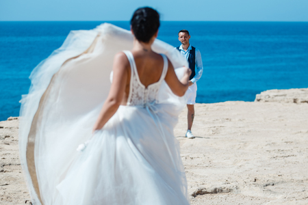 Beautiful smiling young bride and groom walking on the beach, kissing and having fun, wedding ceremony near the rocks and sea. Wedding ceremony on coast of Cyprusの写真素材