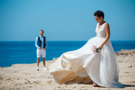 Beautiful smiling young bride and groom walking on the beach, kissing and having fun, wedding ceremony near the rocks and sea. Wedding ceremony on coast of Cyprusの写真素材