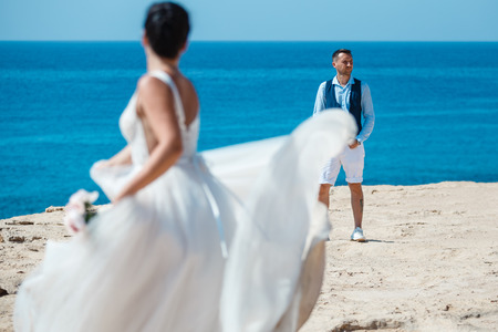 Beautiful smiling young bride and groom walking on the beach, kissing and having fun, wedding ceremony near the rocks and sea. Wedding ceremony on coast of Cyprusの写真素材