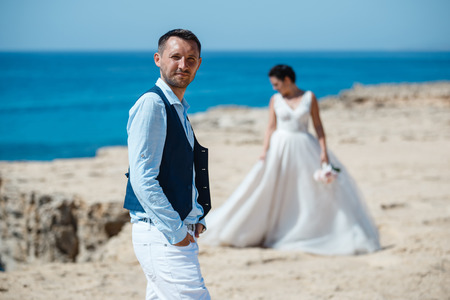 Beautiful smiling young bride and groom walking on the beach, kissing and having fun, wedding ceremony near the rocks and sea. Wedding ceremony on coast of Cyprusの写真素材