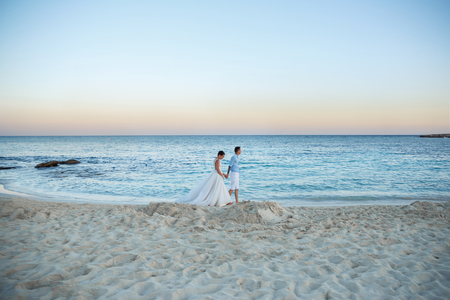 Beautiful smiling young bride and groom walking on the beach, kissing and having fun, wedding ceremony near the rocks and sea. Wedding ceremony on coast of Cyprusの写真素材