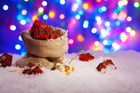 Sack Santa Claus with presents on snow, on a background of colored lights.の写真素材