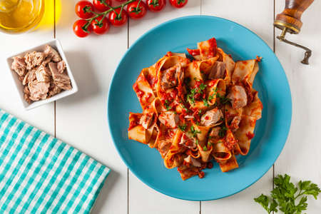 Pappardelle pasta with tuna, in tomato sauce. Served on a blue plate, white boards in the background. Top view.の写真素材