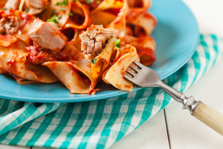 Pappardelle pasta with tuna, in tomato sauce. Served on a blue plate, white boards in the background. Front view. Close up.の写真素材
