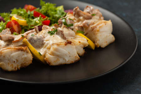 Fried cod served with mushroom sauce and salads. Portion on a black plate. Dark background.