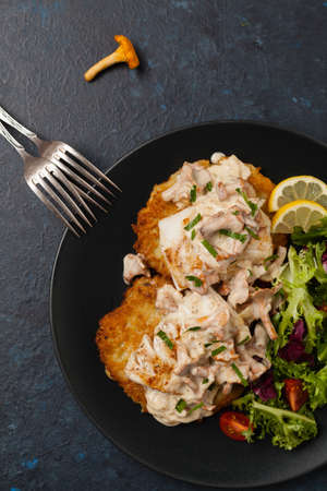 Fried cod, served on potato pancakes with mushrooms sauce and salads. Portion on a black plate. Dark background.の写真素材