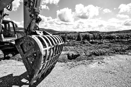 Excavator bucket on the asphalt road near the vineyard. Excavator in construction site on the Italian wine farm surrounded with vineyards background. Black and white pictureの写真素材