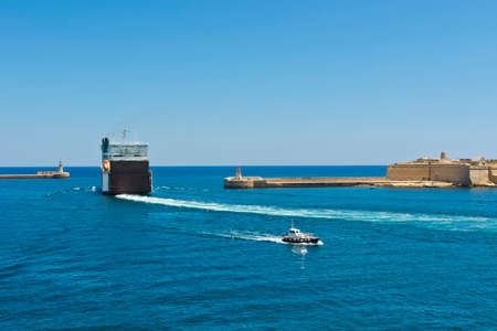 Cargo ship leaves the harbor of Valletta. Lighthouses indicate the entrance to the ports of Maltaの写真素材