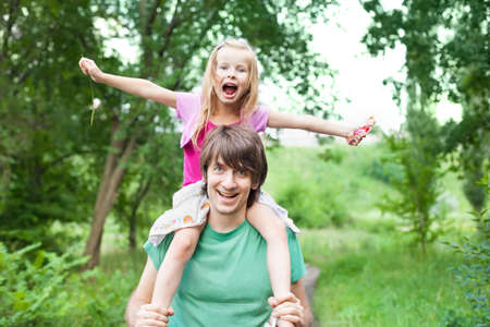 father and daughter.Young man holding blonde little girl on shoulders in park in summerの写真素材
