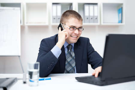 Portrait of smiling young business man talking on cell phone and working on a laptop at officeの写真素材