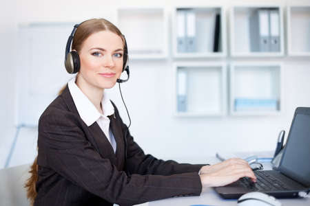 Portrait of pretty young female operator sitting at office desk with headsetの写真素材