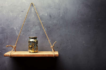Glass jar with coins on the old wood shelf hanging on rope on textured wall background with copy...