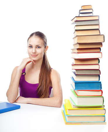 Portrait of pretty young smiling student girl sitting at the table with high stack of books isolated on white backgroundの写真素材