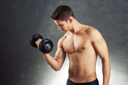 Portrait of young bodybuilder man exercising with dumbbell for training his biceps against gray textured backgroundの写真素材
