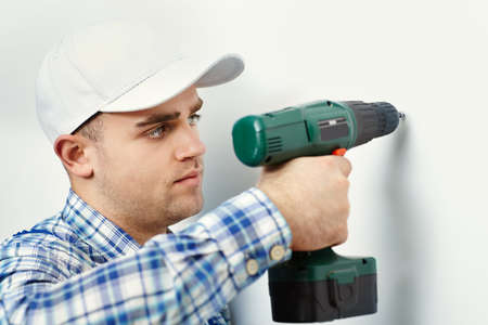 Young worker man with electric drill making hole in white wallの写真素材