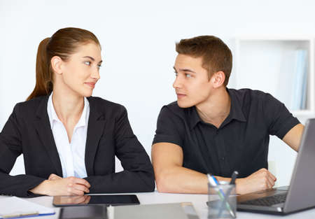 Two young happy businesspeople sitting at table in office and looking at each otherの写真素材