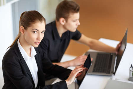 Two beautiful young businesspeople sitting at table in office and working at laptop and tablet pcの写真素材