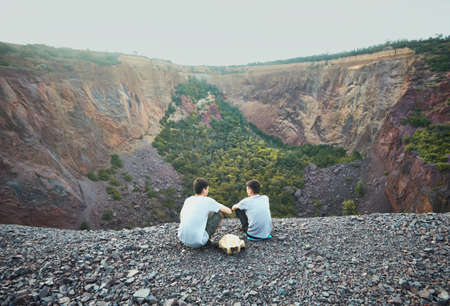 Two tourist young men sitting on rocky cliff and admiring view of abandoned mineの写真素材