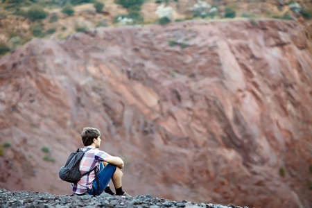 Young tourist man with backpack sitting on rocky cliff and enjoying beautiful viewの写真素材