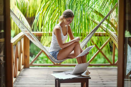 Young beautiful woman sitting in a hammock with documents and laptop in a tropical resort.の写真素材