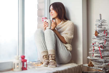 Happy beautiful woman drinking hot coffee sitting on window sill in christmas decorated home. Holiday conceptの写真素材