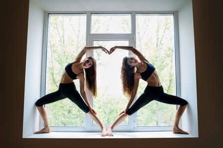 Two young beautiful women doing yoga asana showing heart symbol on window sillの写真素材