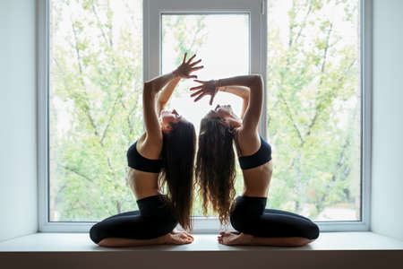 Two young beautiful women doing yoga asana on window sillの写真素材