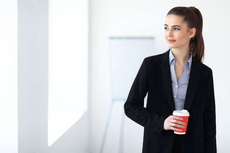 Portrait of young beautiful business woman with cup of coffee in the office backgroundの写真素材