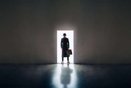 Man silhouette in hat and raincoat standing in the light of opening door in dark room