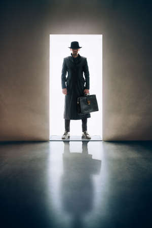 Man silhouette in hat and raincoat standing in the light of opening door in dark roomの写真素材